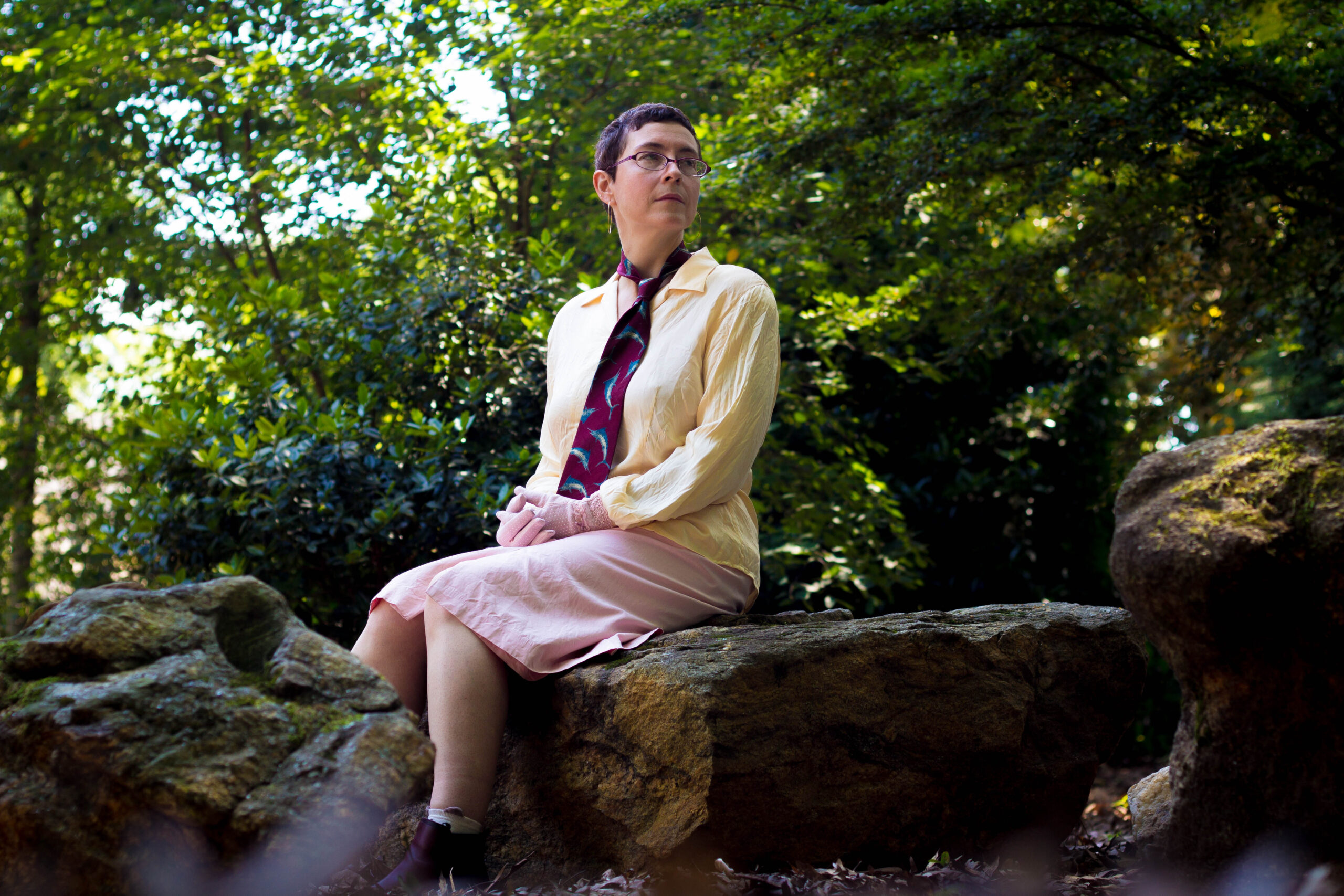 An artist sitting in soft light surrounded by trees, with a serene smile. She is wearing pink gloves and a tie with swordfish on it. Located in Athens GA.