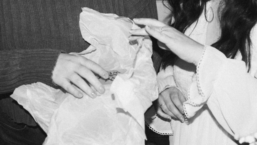Close up of two sets of hands unwrapping a gift, an engagement right is visible on one hand. A black and white film photo from an engagement party.