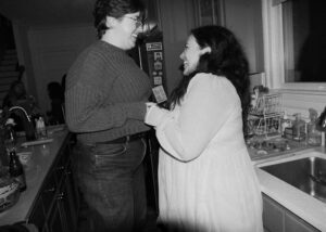 A queer couple holds hands and jumps with joy during their engagement party
