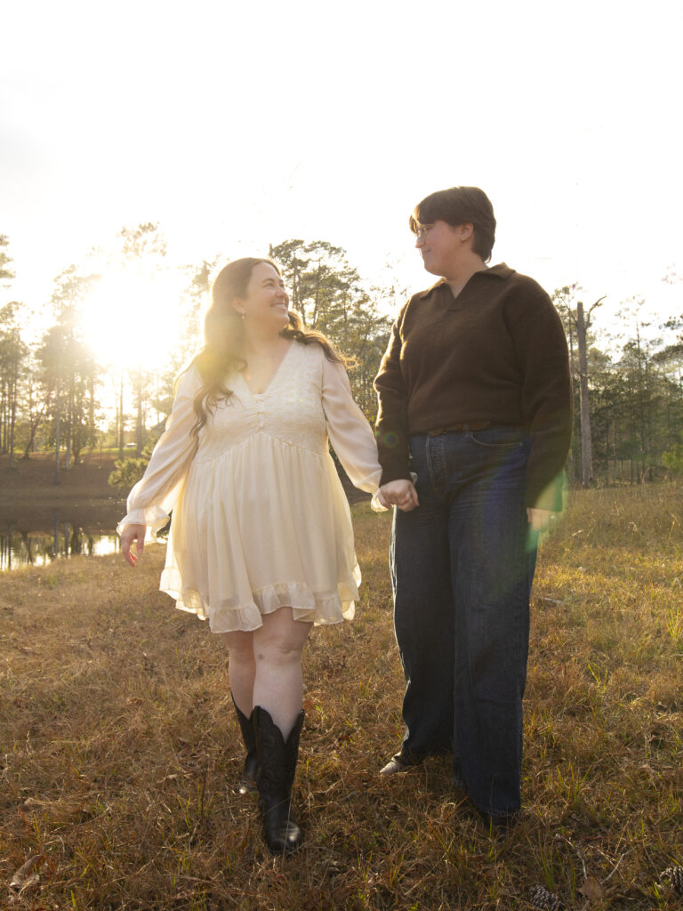 LGBTQ+ couple walks, holding hands, by a lake. The sunset flares soft golden like across the image. 