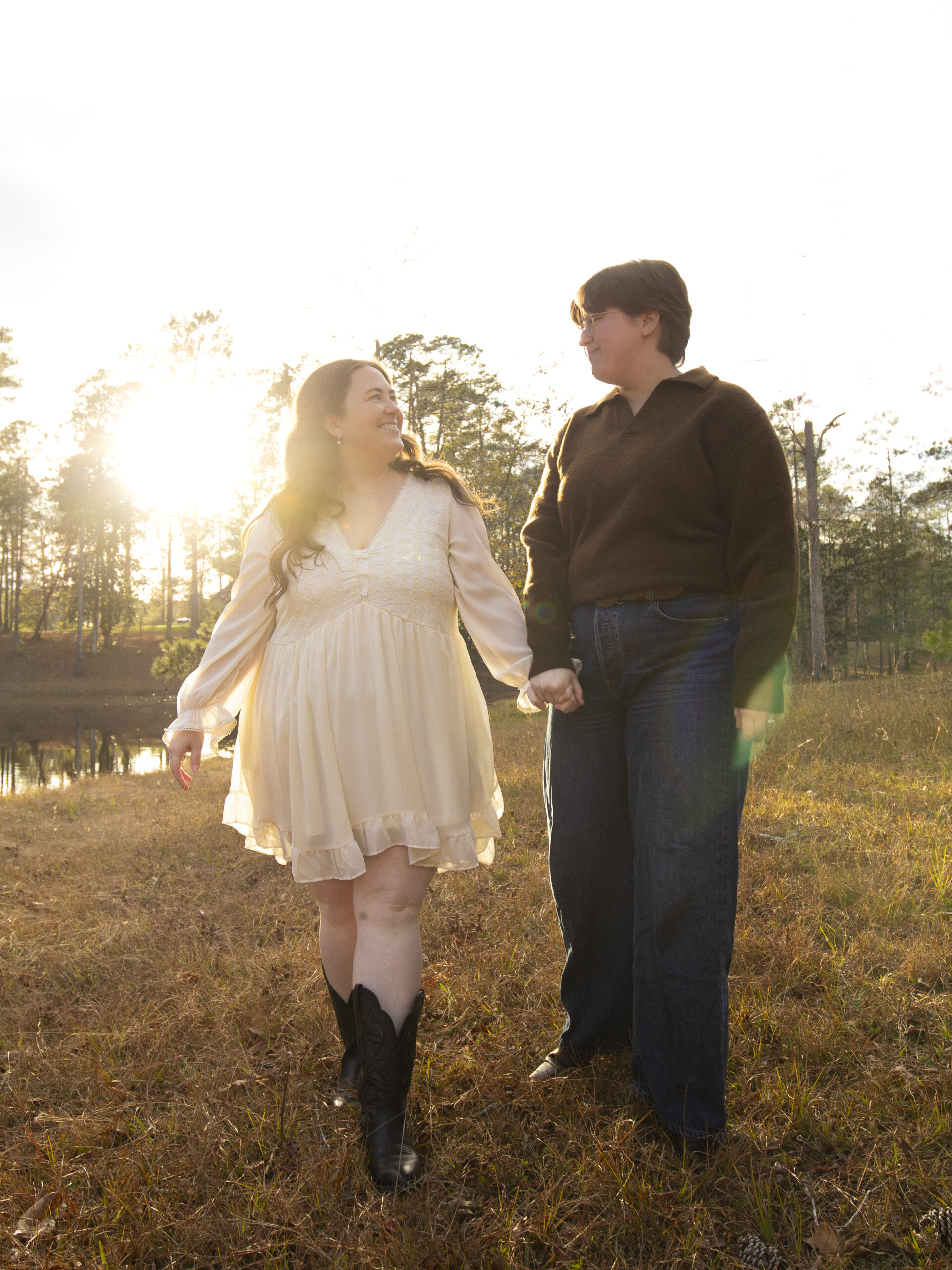 LGBTQ+ couple walks, holding hands, by a lake. The sunset flares soft golden like across the image.
