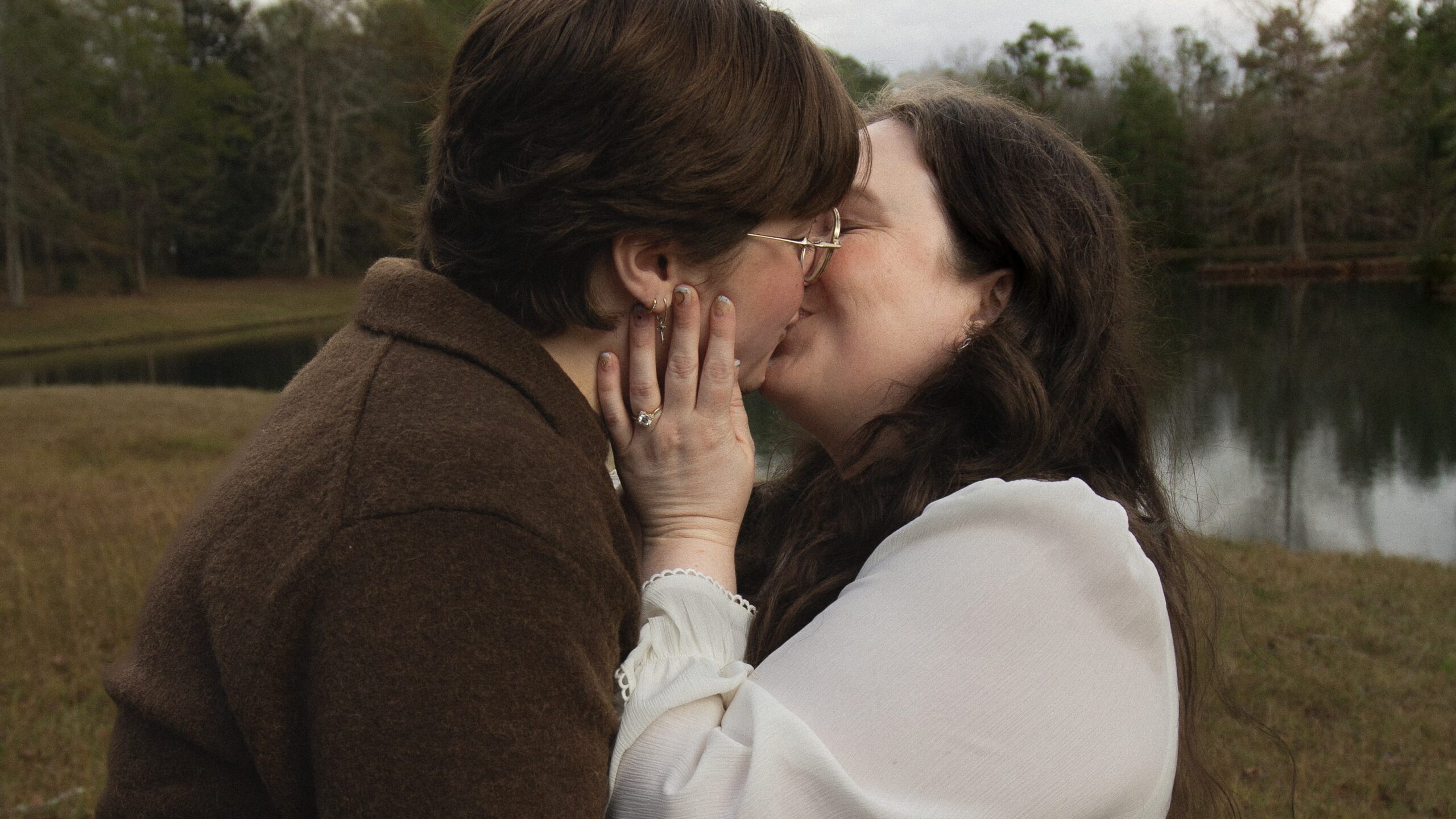 Kissing by a foggy lake, a new engagement right is prominent on one woman's hand