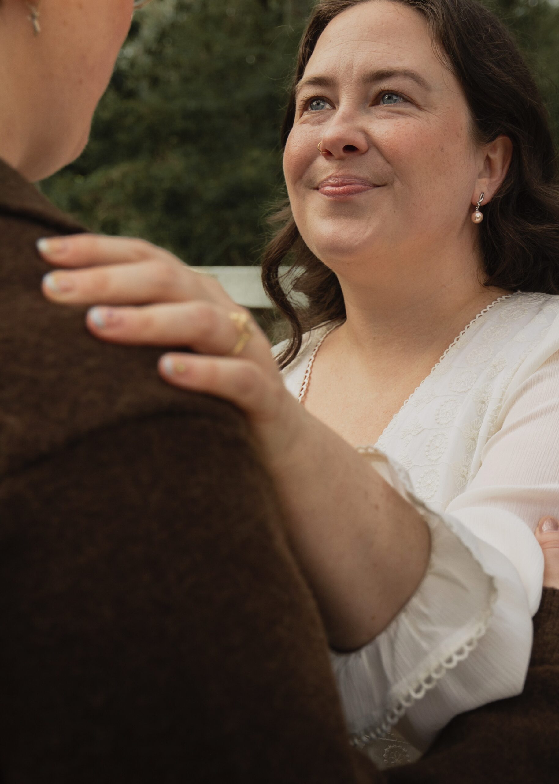 A new engagement ring is prominent, a woman has her left hand on her partner's shoulder and is smiling softly with love