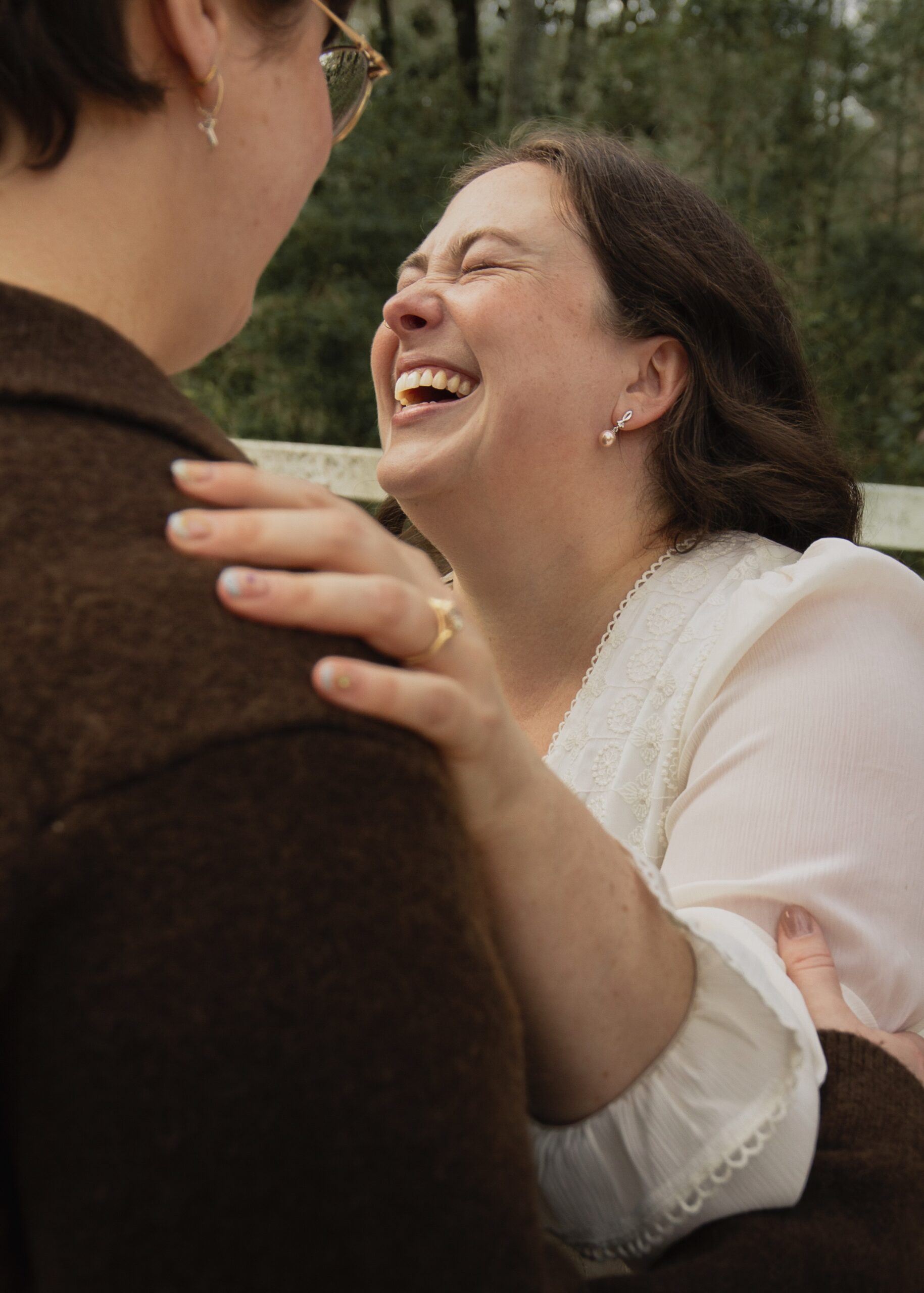 A new engagement ring is prominent, a woman has her left hand on her partner's shoulder and is laughing