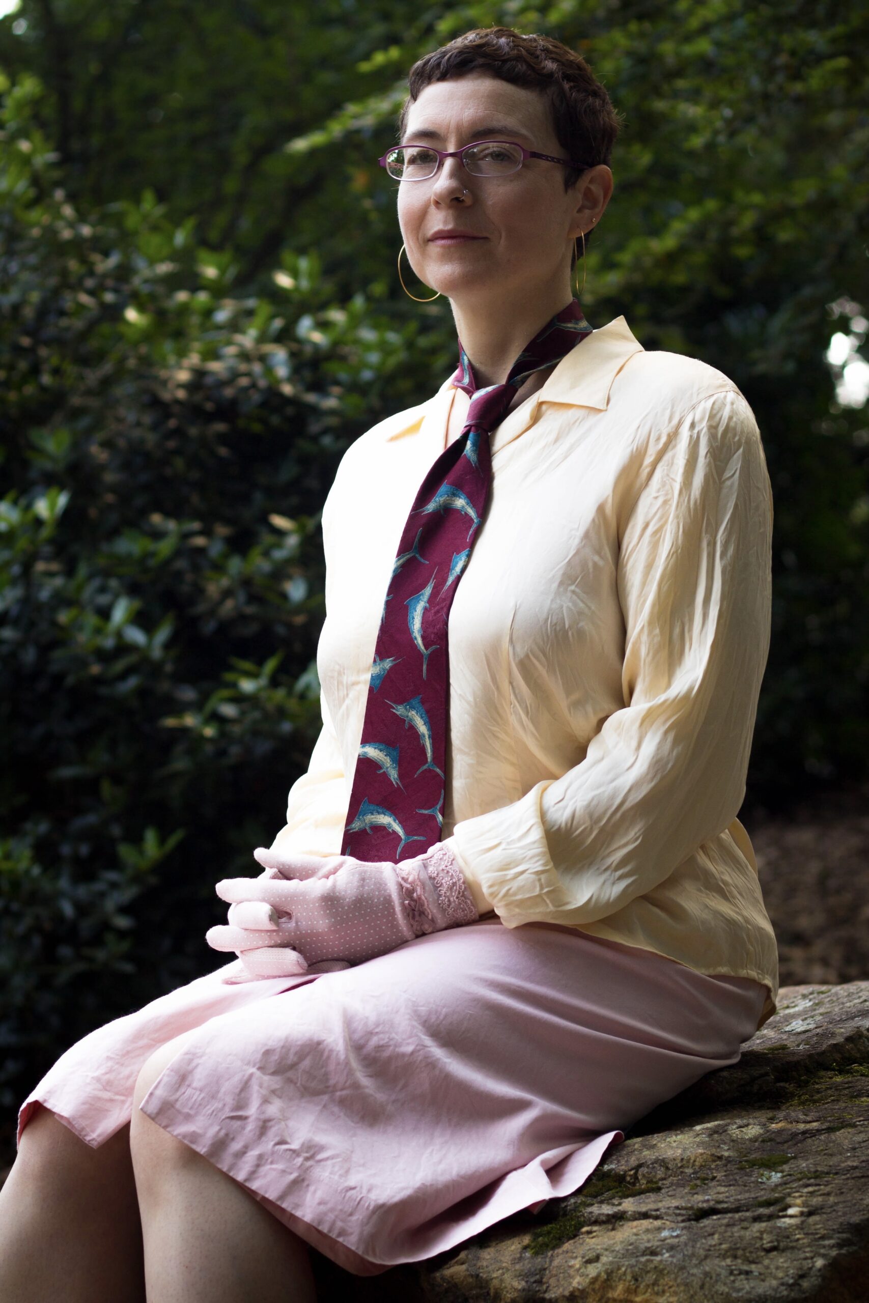 An artist sitting in soft light surrounded by trees, with a serene smile. She is wearing pink gloves and a tie with swordfish on it. Located in Athens GA.